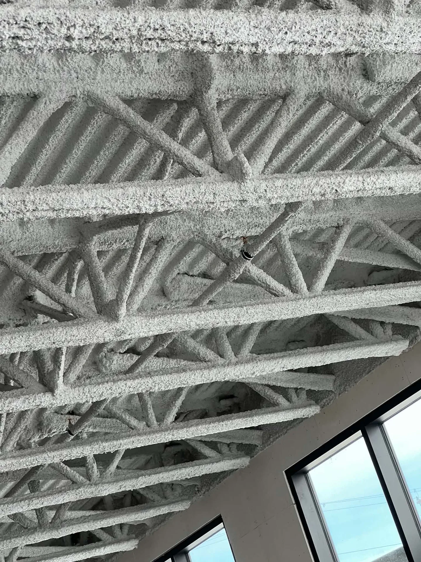 Fireproofing covering ceiling trusses beside large windows.